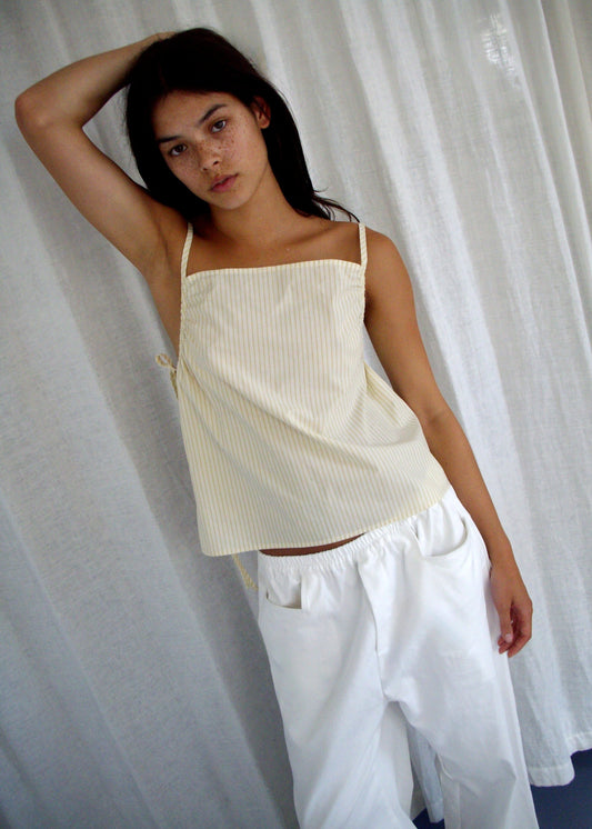 A person with long dark hair stands before white curtains, wearing the Gathered Strap Top - Butter Stripe from Deiji Studios and loose white pants, posing with one arm raised and leaning to the side.