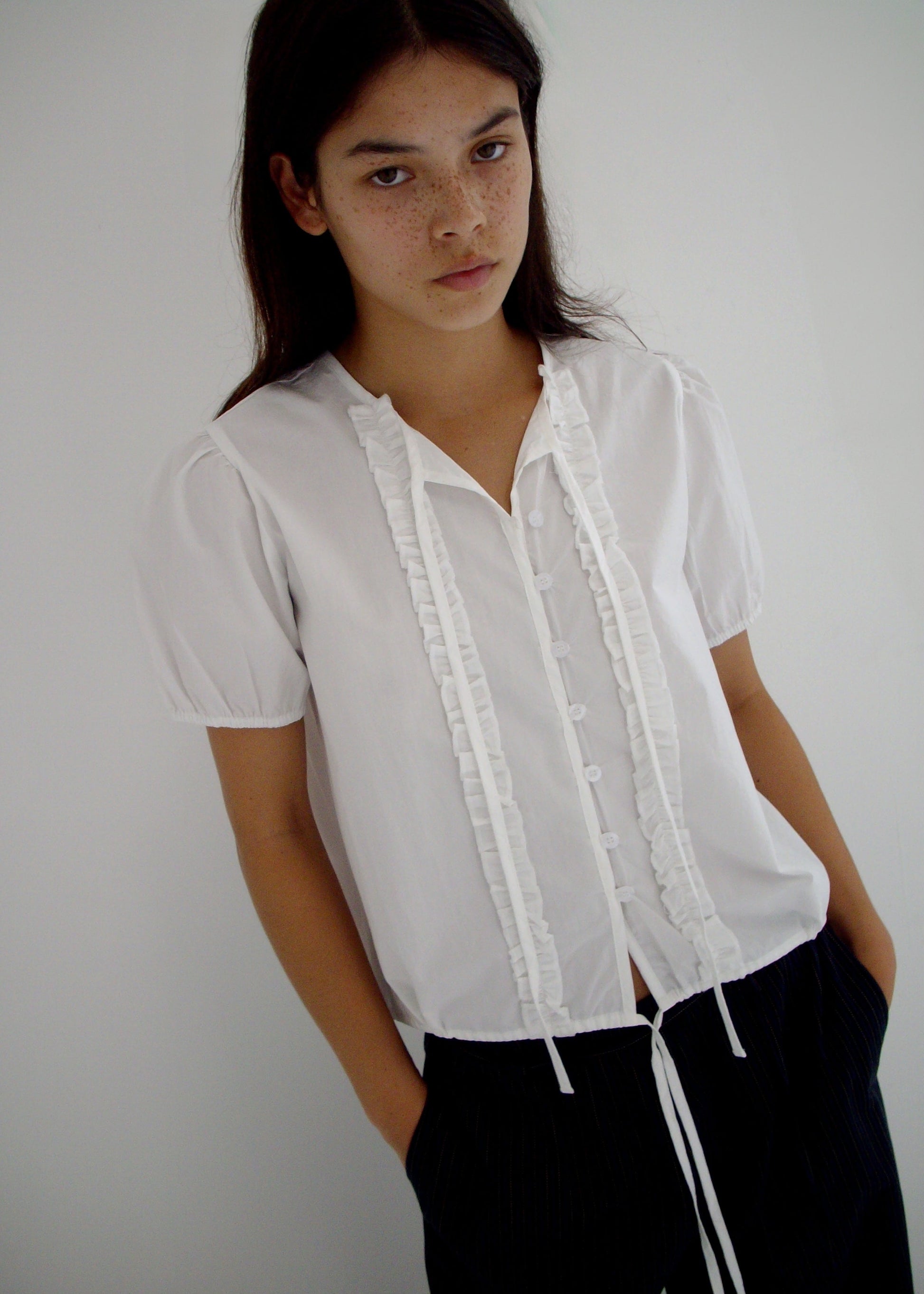 A person with long dark hair and freckles stands against a white backdrop, wearing the 0 Double Frill Top - White and black pants, hands in pockets and a neutral expression.