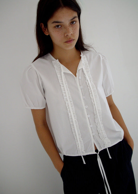 A person with long dark hair and freckles stands against a white backdrop, wearing the 0 Double Frill Top - White and black pants, hands in pockets and a neutral expression.