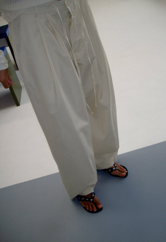 female model wears the turn pant in off white, a relaxed tailored pant features soft pleating at the front, buttoned waistband and adjustable inner drawcord. designed with hidden side seam pockets and pockets at the back, made from 100% GOTS certified organic cotton paired with a white top