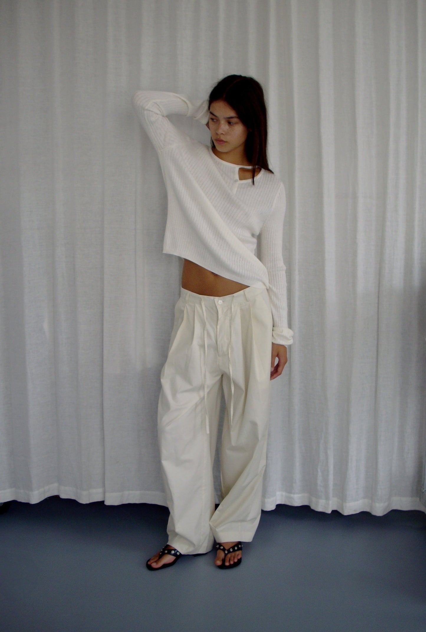 female model wears the turn pant in off white, a relaxed tailored pant features soft pleating at the front, buttoned waistband and adjustable inner drawcord. designed with hidden side seam pockets and pockets at the back, made from 100% GOTS certified organic cotton paired with a white top