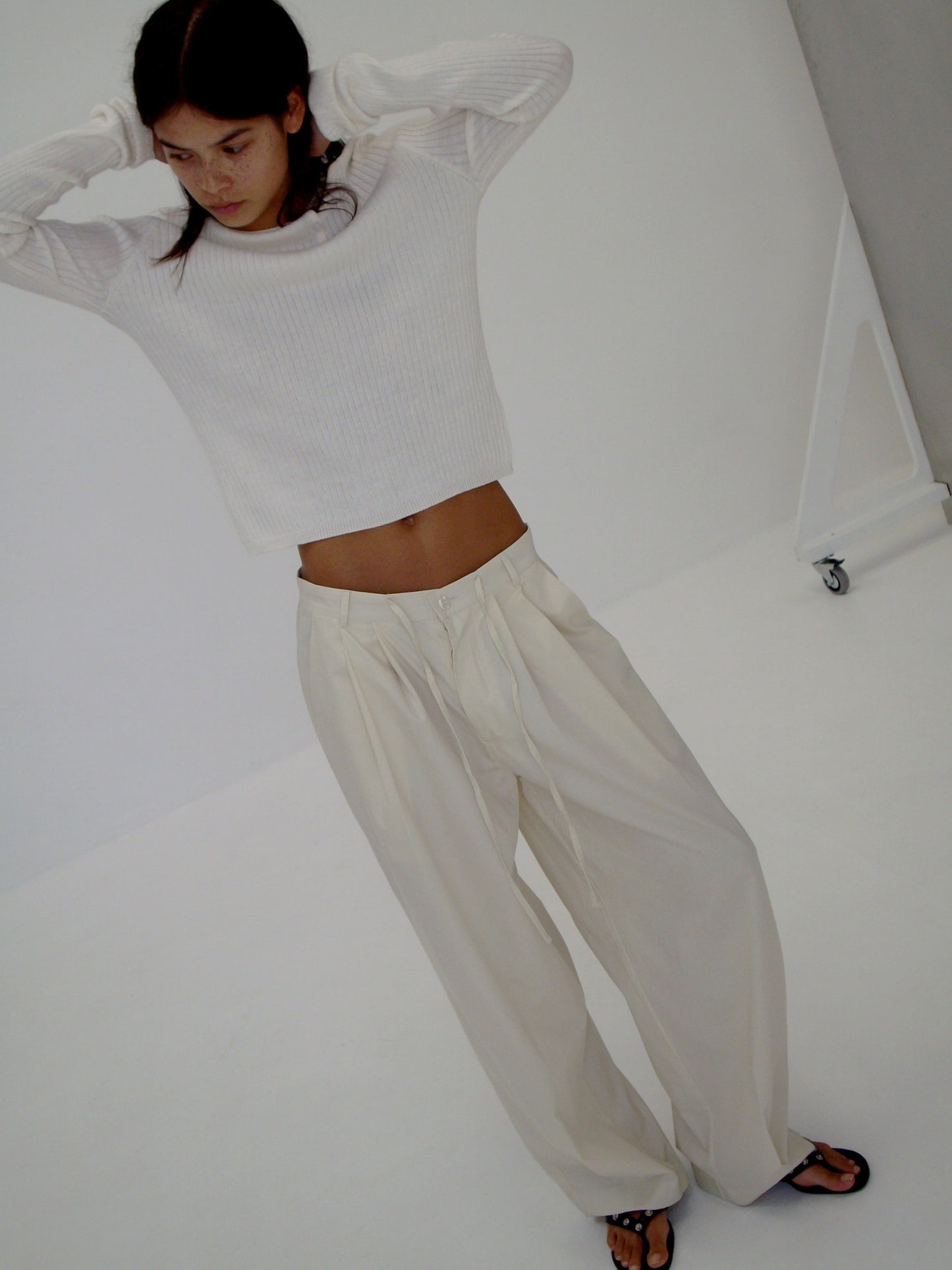female model wears the turn pant in off white, a relaxed tailored pant features soft pleating at the front, buttoned waistband and adjustable inner drawcord. designed with hidden side seam pockets and pockets at the back, made from 100% GOTS certified organic cotton paired with a white top