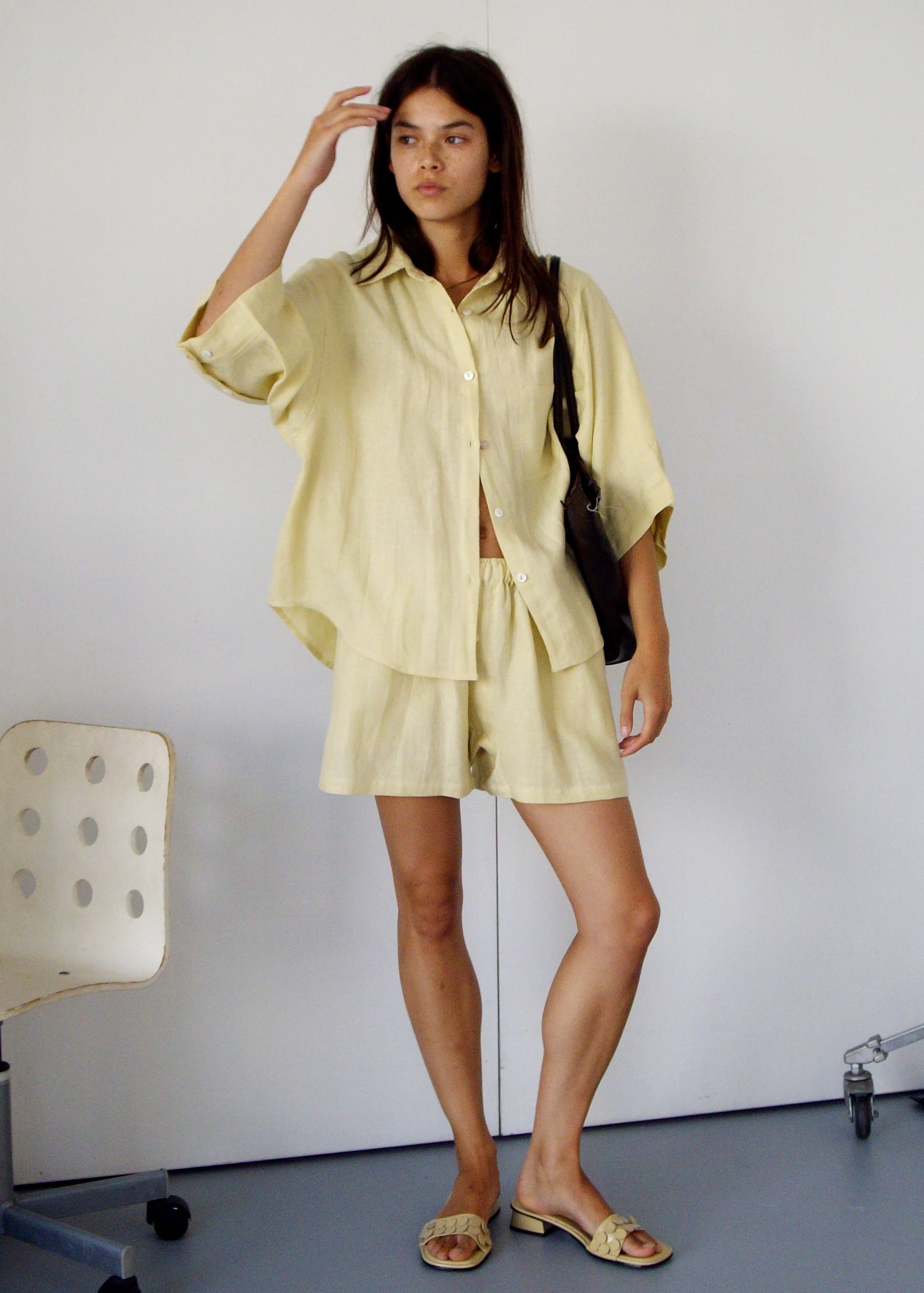 A woman with shoulder-length dark hair stands indoors, wearing the 03 Set - Butter Yellow. She carries a black bag, wears sandals, and is near a white chair and part of a wheeled stand.