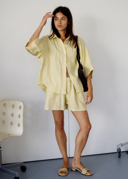 A woman with shoulder-length dark hair stands indoors, wearing the 03 Set - Butter Yellow. She carries a black bag, wears sandals, and is near a white chair and part of a wheeled stand.