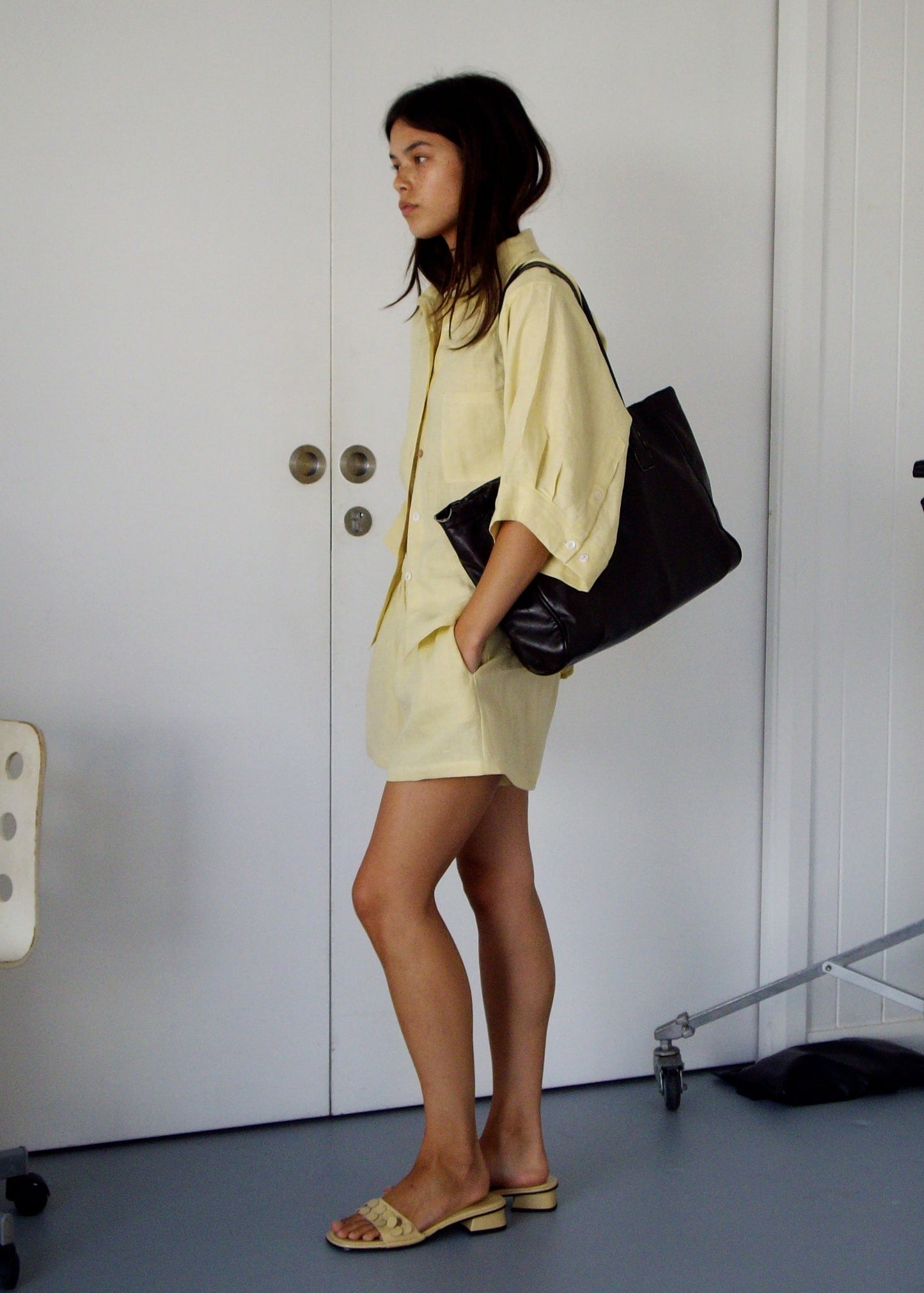 A woman stands indoors wearing the 03 Set - Butter Yellow, beige sandals, and carrying a large black bag. She faces left beside a white door and an office chair.