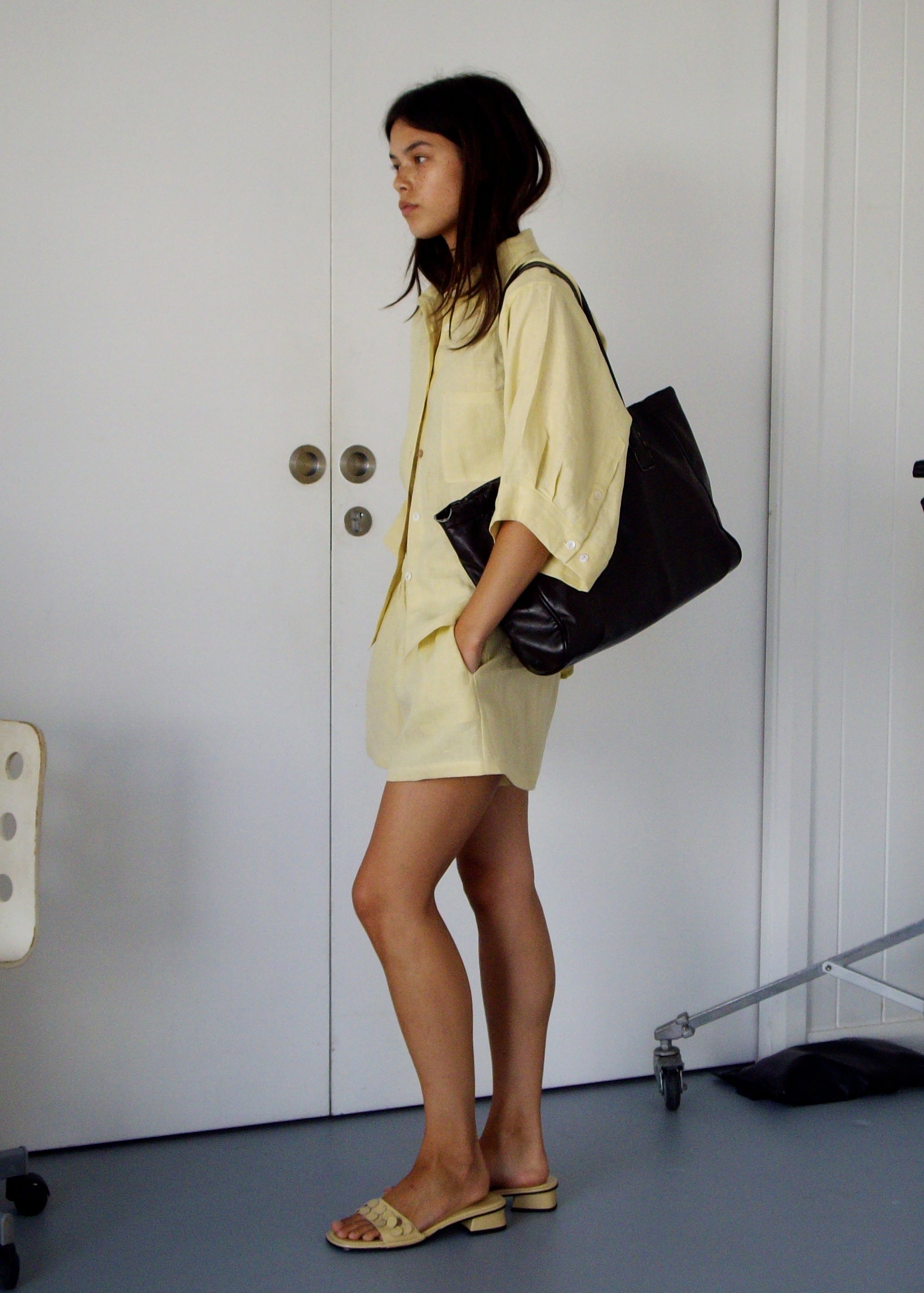 A woman stands indoors wearing the 03 Set - Butter Yellow, beige sandals, and carrying a large black bag. She faces left beside a white door and an office chair.