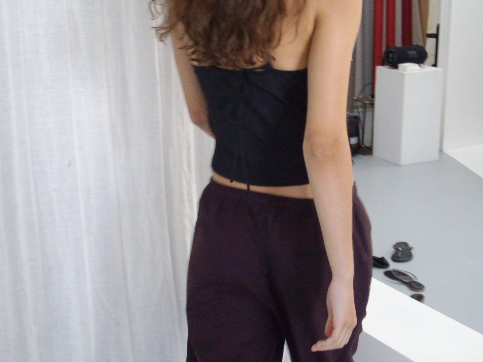 A person with wavy brown hair, wearing a black top and the Drawcord Culotte - Burgundy Brown Stripe by Deiji Studios, stands indoors facing away from the camera, surrounded by white curtains, a white platform, and scattered objects on the floor.