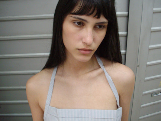 A close-up of a woman with dark hair and a fringe outdoors, wearing the Folded Top - Cloud Stripe by Deiji Studios, a pale grey-blue fine-striped fold-over halter top.