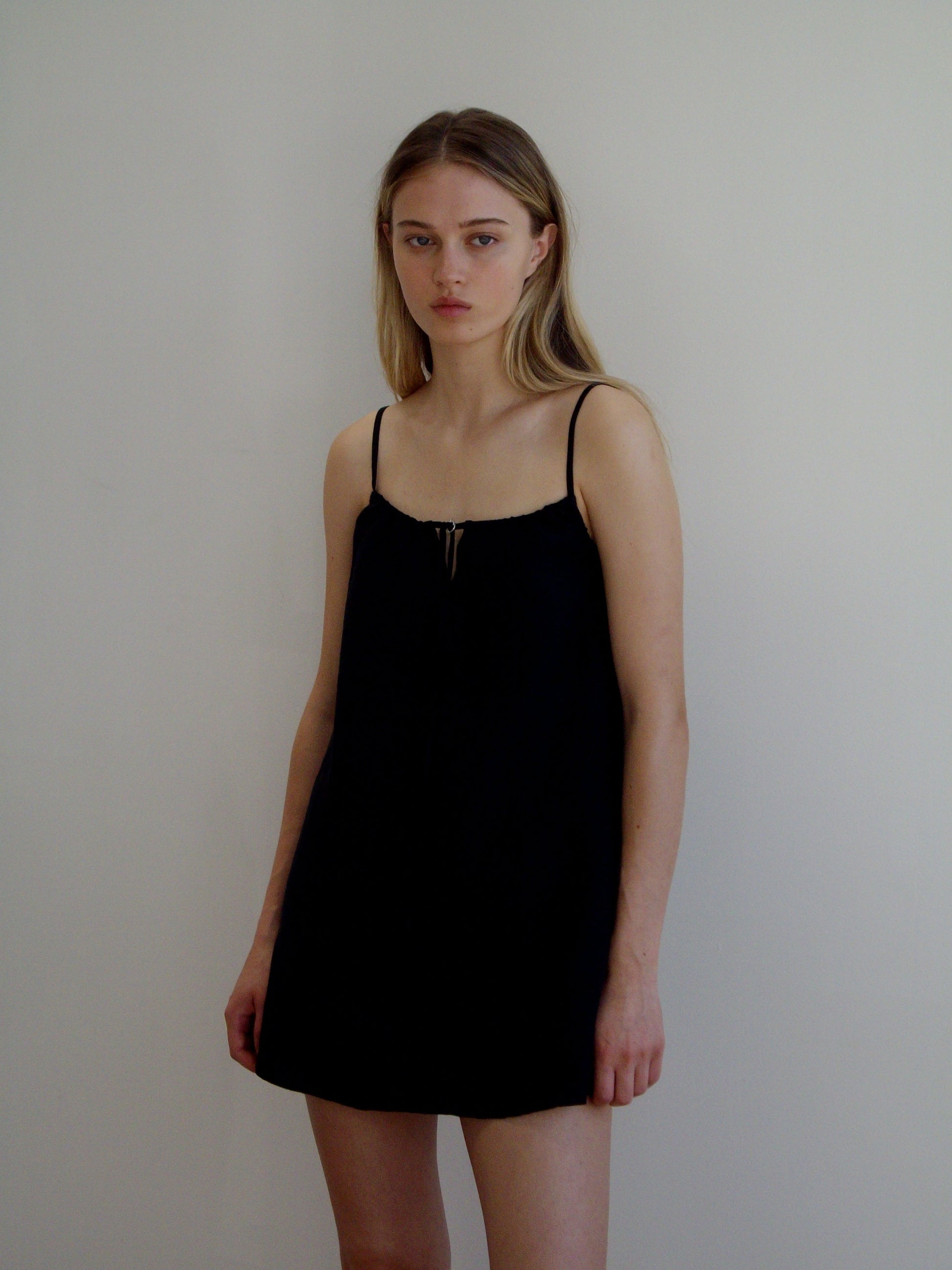 A woman with long blonde hair stands against a plain white wall, wearing the Lettuce Mini Dress - Black by Deiji Studios. She has a neutral expression and her arms are relaxed at her sides.