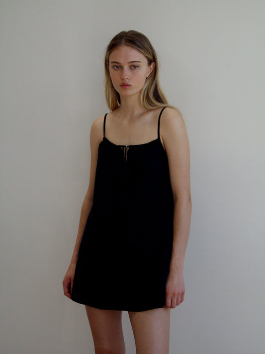A woman with long blonde hair stands against a plain white wall, wearing the Lettuce Mini Dress - Black by Deiji Studios. She has a neutral expression and her arms are relaxed at her sides.