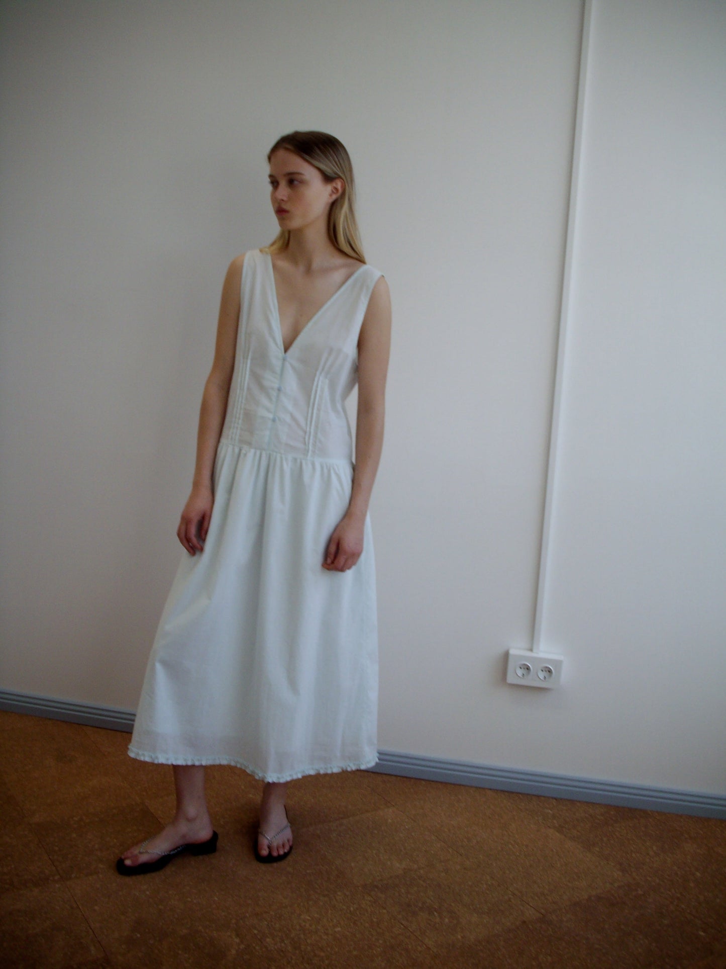 A woman stands indoors against a plain wall, wearing a sleeveless mint Pintuck Dress from Deiji Studios with a deep V-neck and flat black sandals. She has straight, light brown hair and gazes slightly to the side.