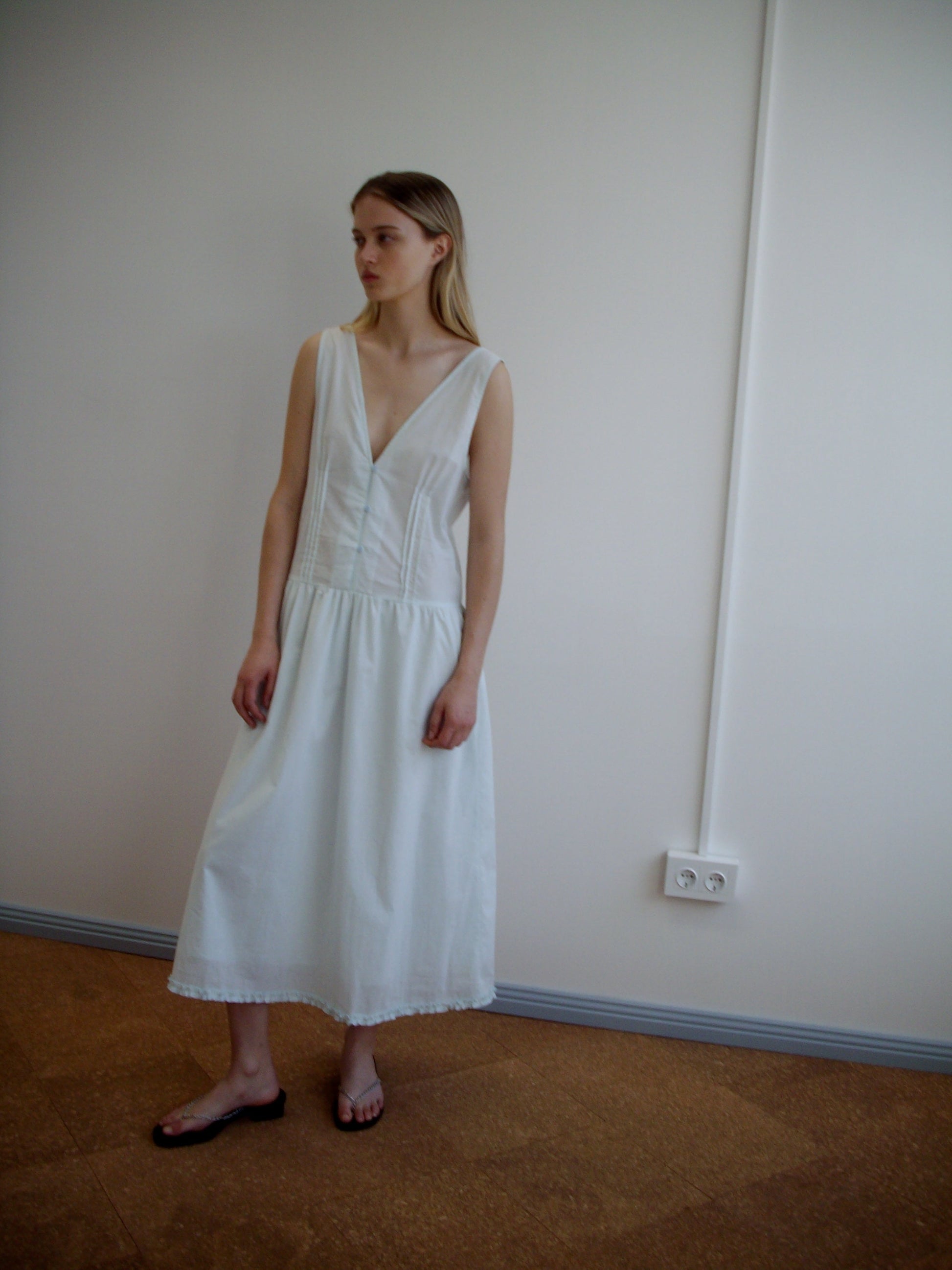 A woman stands indoors against a plain wall, wearing a sleeveless mint Pintuck Dress from Deiji Studios with a deep V-neck and flat black sandals. She has straight, light brown hair and gazes slightly to the side.