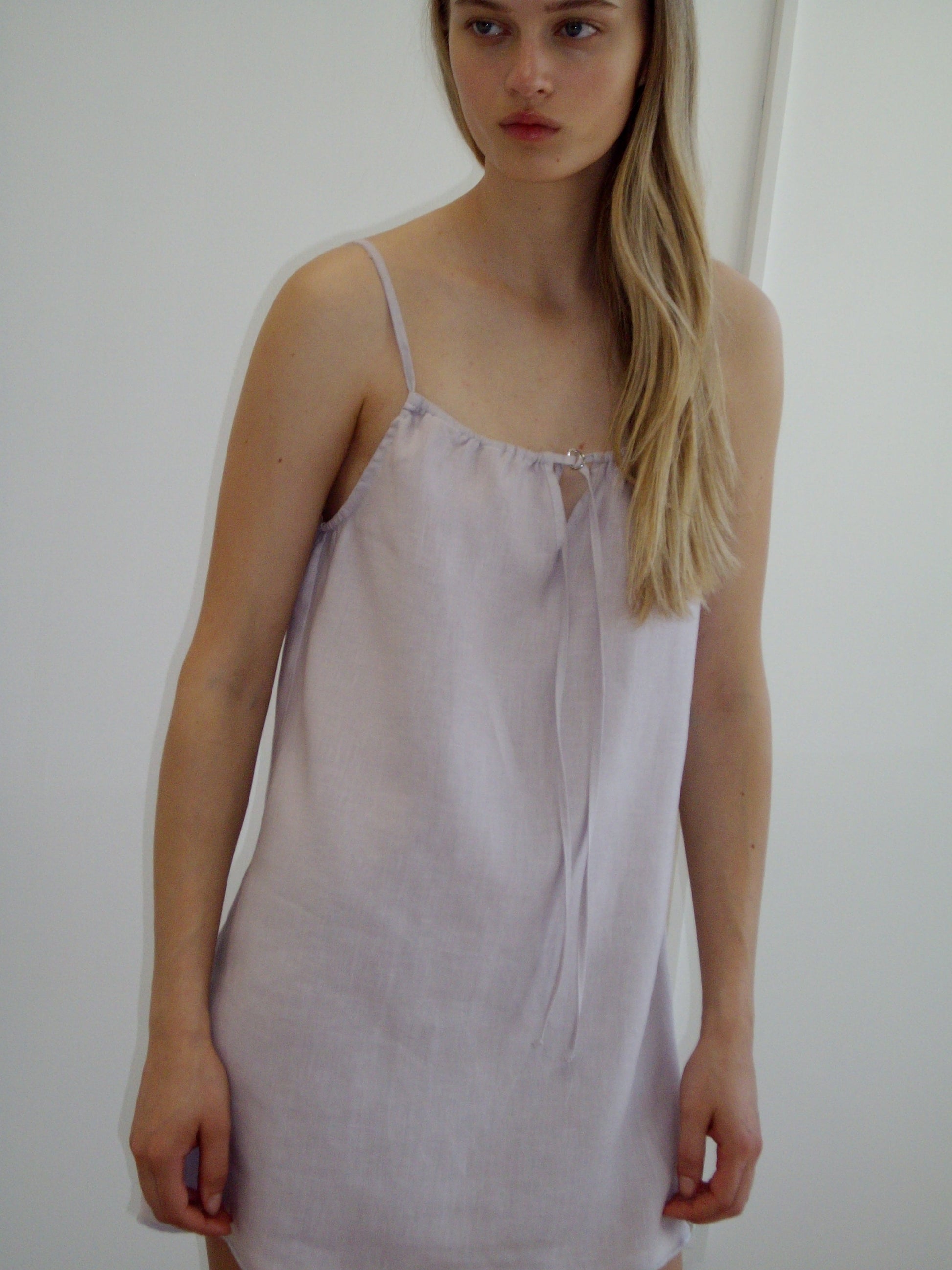 A woman with long blonde hair models the Lettuce Mini Dress, a light lavender sleeveless style with thin straps, standing against a plain white background.