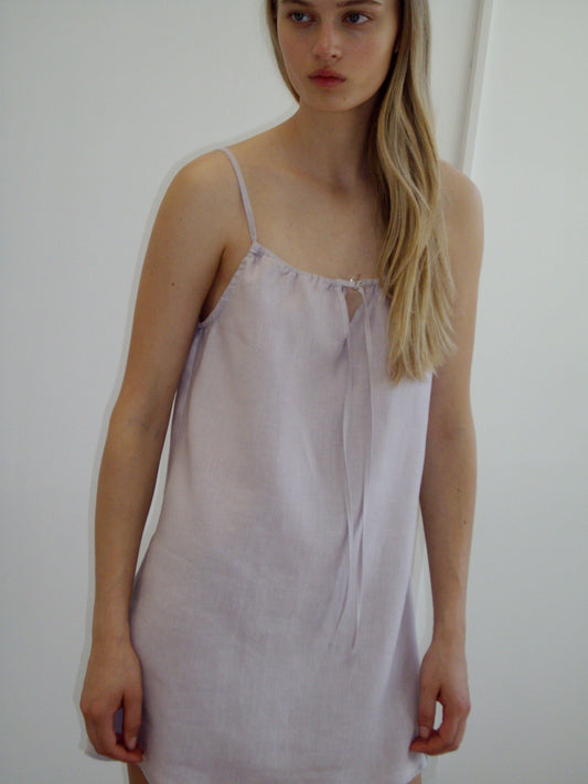 A woman with long blonde hair models the Lettuce Mini Dress, a light lavender sleeveless style with thin straps, standing against a plain white background.