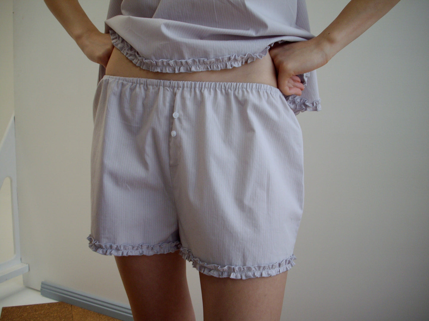 A person models the Yoke Set - Lilac Stripe by Deiji Studios—light, ruffled pajamas shown from torso to upper legs, standing hands on hips against a neutral background.