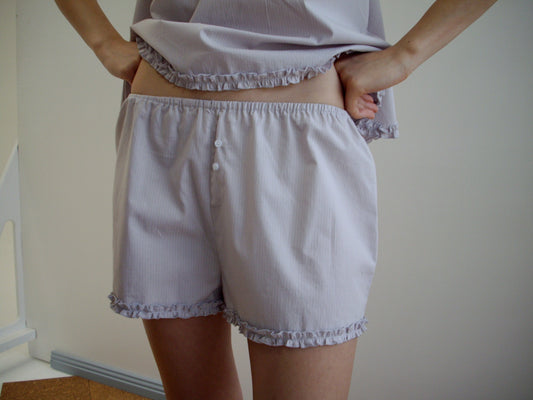 A person models the Yoke Set - Lilac Stripe by Deiji Studios—light, ruffled pajamas shown from torso to upper legs, standing hands on hips against a neutral background.