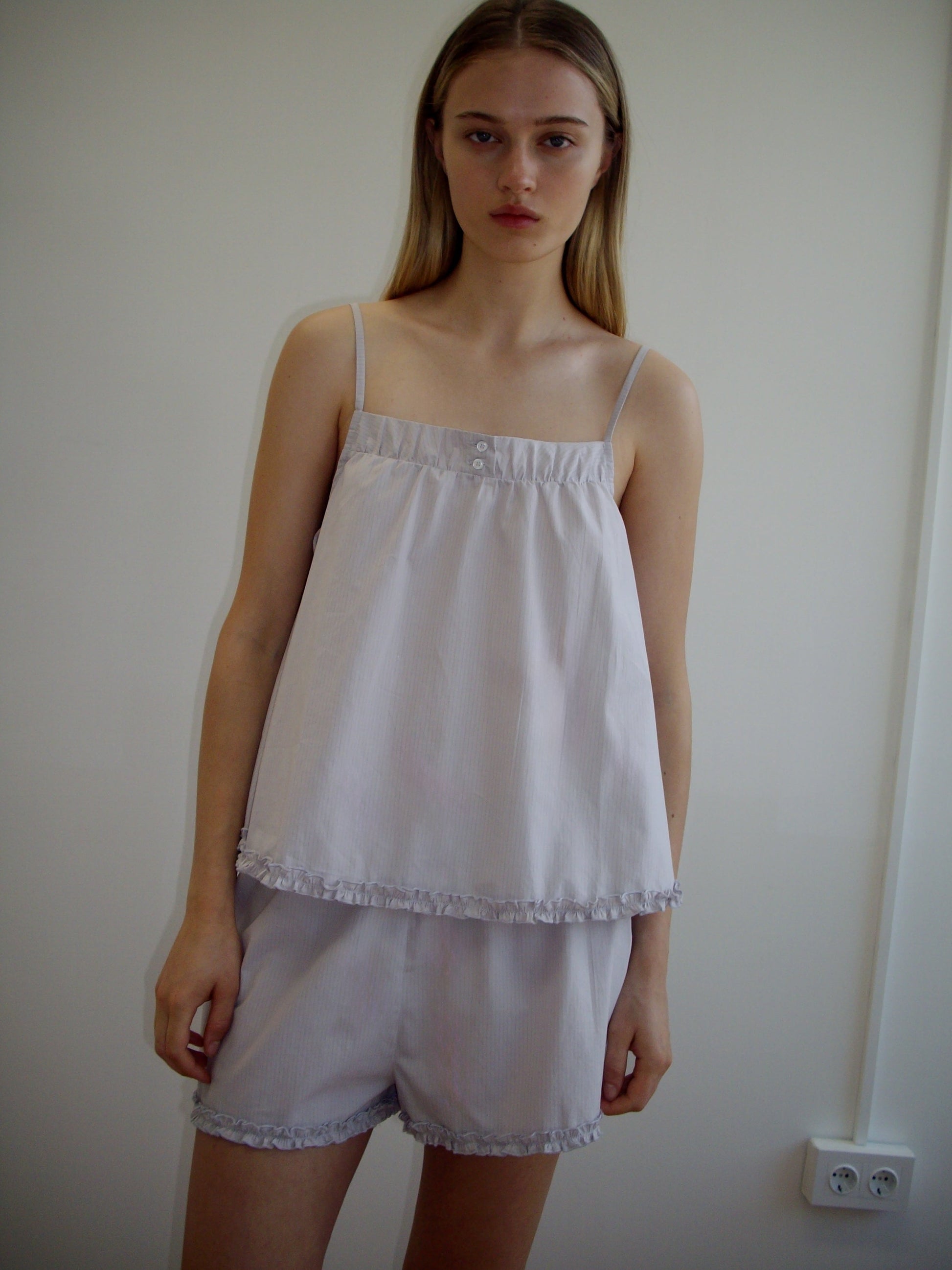A young woman with long, straight blonde hair is wearing the Yoke Set - Lilac Stripe by Deiji Studios, featuring a babydoll top and matching shorts with delicate ruffle trim, while standing indoors against an off-white wall.