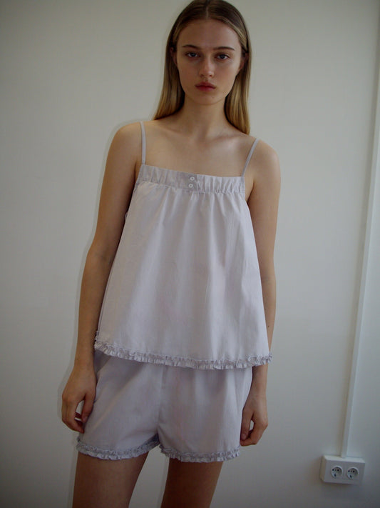 A young woman with long, straight blonde hair is wearing the Yoke Set - Lilac Stripe by Deiji Studios, featuring a babydoll top and matching shorts with delicate ruffle trim, while standing indoors against an off-white wall.