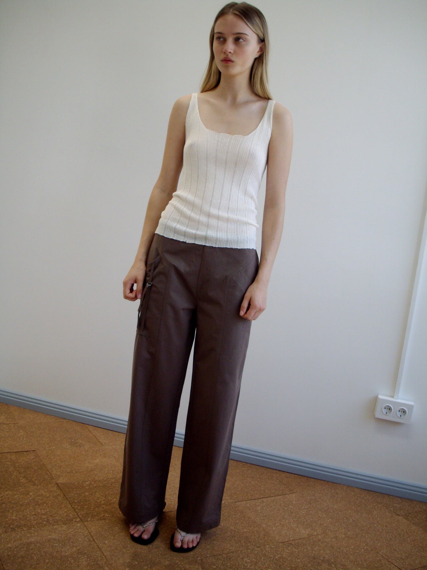 A woman stands indoors against a plain wall wearing the Open Work Tank - White by Deiji Studios, wide-legged brown ecoviscose linen pants, and dark sandals. She has long straight hair and a neutral expression.
