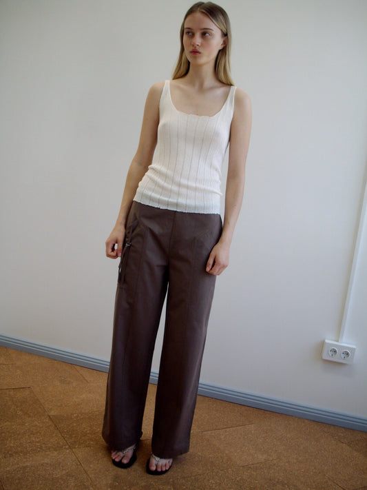 A woman stands indoors against a plain wall wearing the Open Work Tank - White by Deiji Studios, wide-legged brown ecoviscose linen pants, and dark sandals. She has long straight hair and a neutral expression.