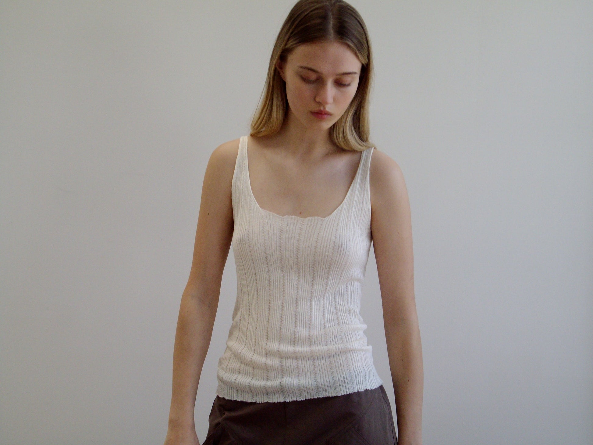 A young woman with long, straight, light brown hair stands against a plain white background, wearing the Open Work Tank - White by Deiji Studios and brown ecoviscose linen pants, looking downward with a neutral expression.