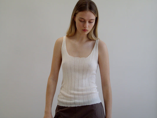 A young woman with long, straight, light brown hair stands against a plain white background, wearing the Open Work Tank - White by Deiji Studios and brown ecoviscose linen pants, looking downward with a neutral expression.