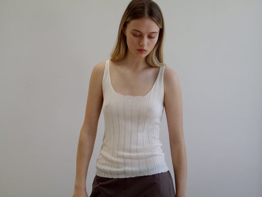 A young woman with long, straight, light brown hair stands against a plain white background, wearing the Open Work Tank - White by Deiji Studios and brown ecoviscose linen pants, looking downward with a neutral expression.