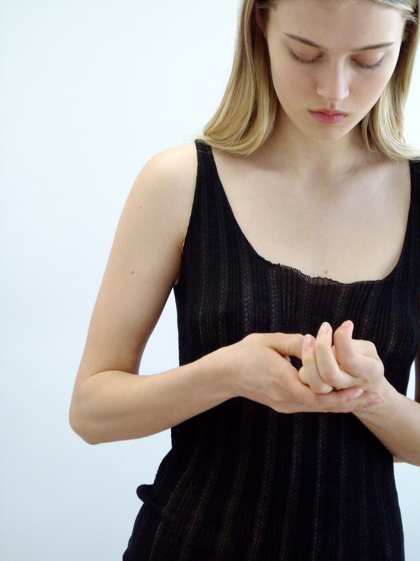 A woman with long blonde hair wears the Open Work Tank - Black by Deiji Studios, gently touching her fingers together while looking down against a plain light background.