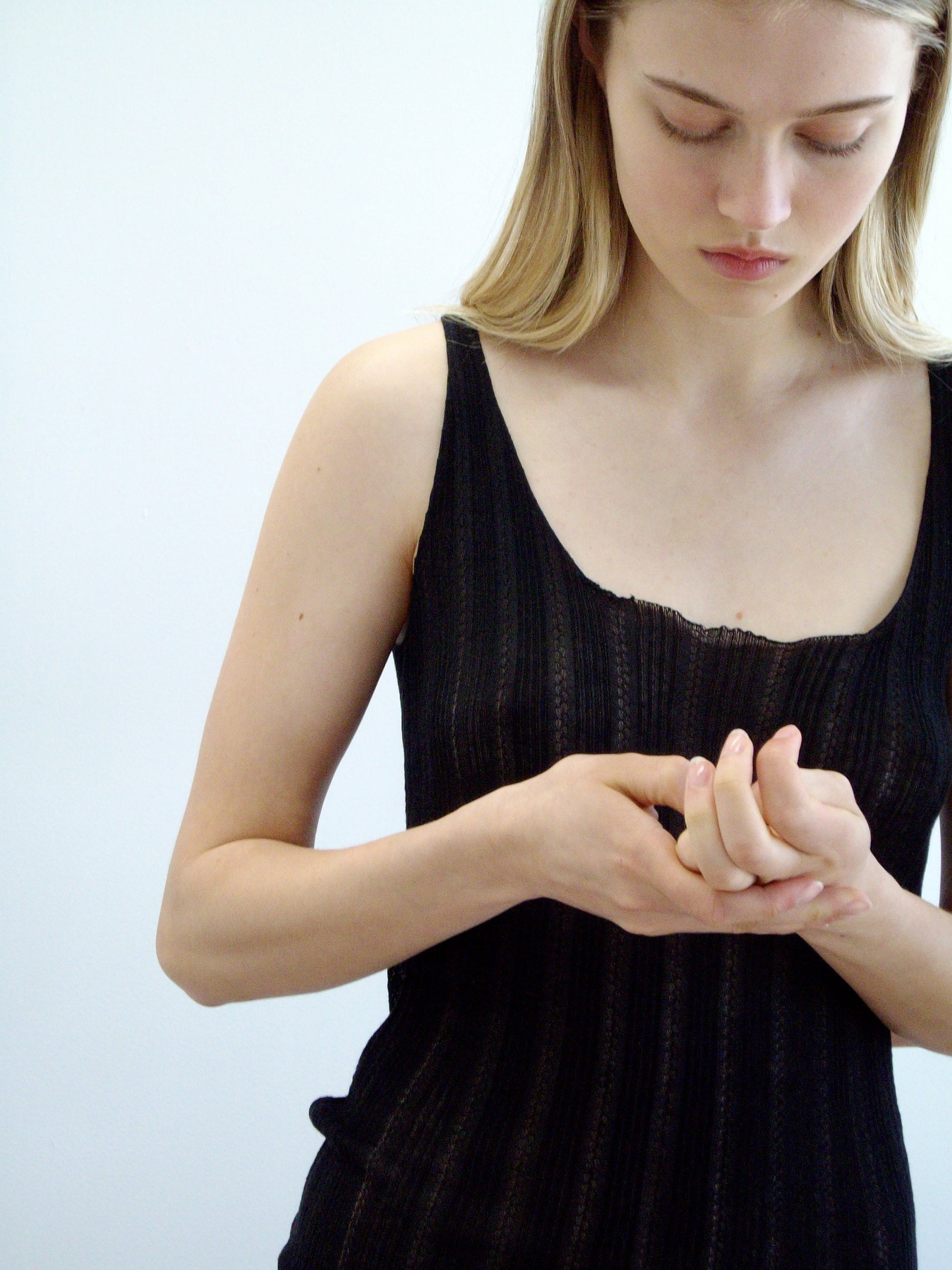 A woman with long blonde hair wears the Open Work Tank - Black by Deiji Studios, gently touching her fingers together while looking down against a plain light background.