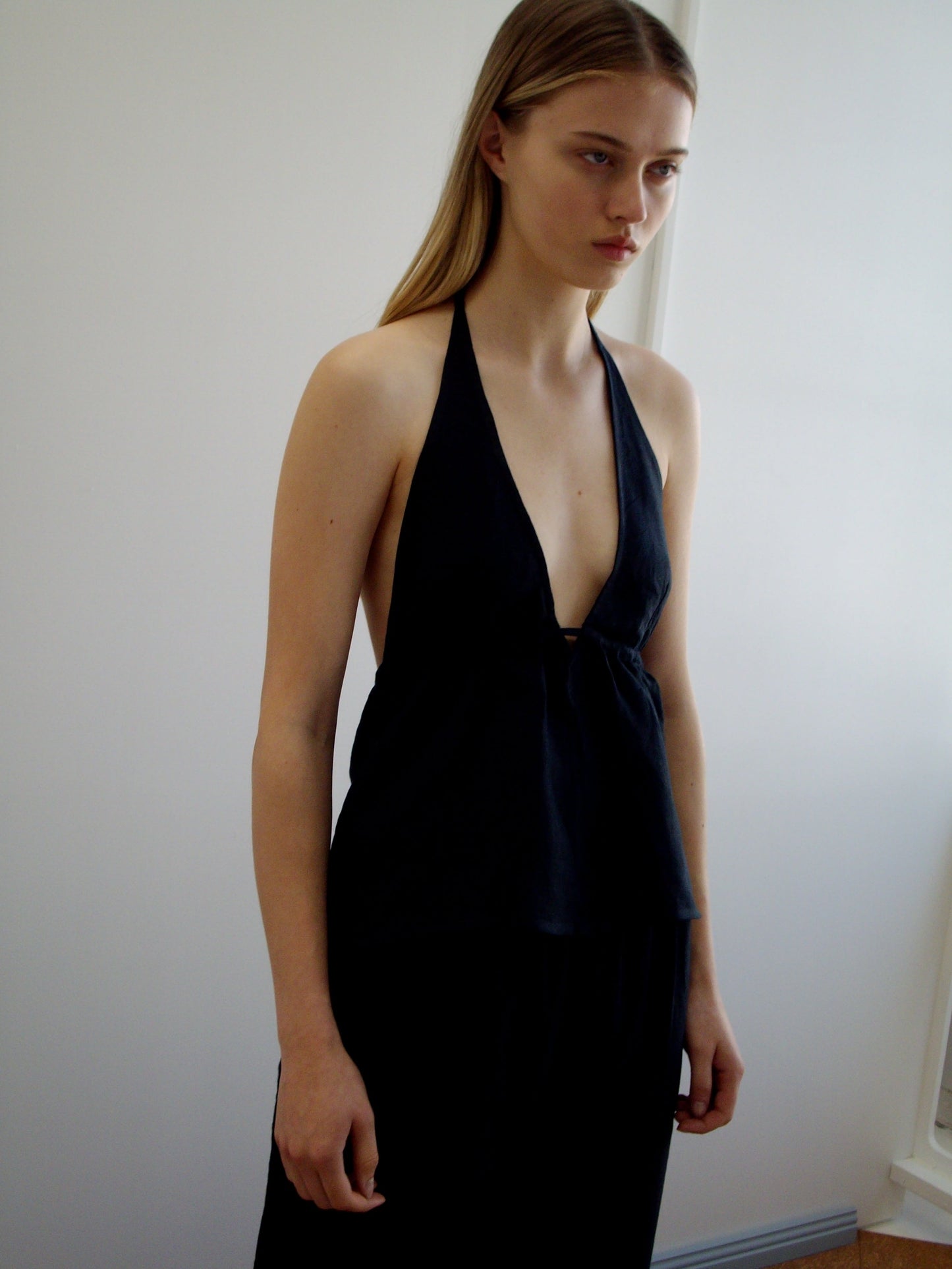 A person with long, straight, light brown hair stands indoors by a white wall, wearing the sleeveless Linen Button Halter top with a deep V-neck. They have a neutral expression and their arms rest relaxed at their sides.