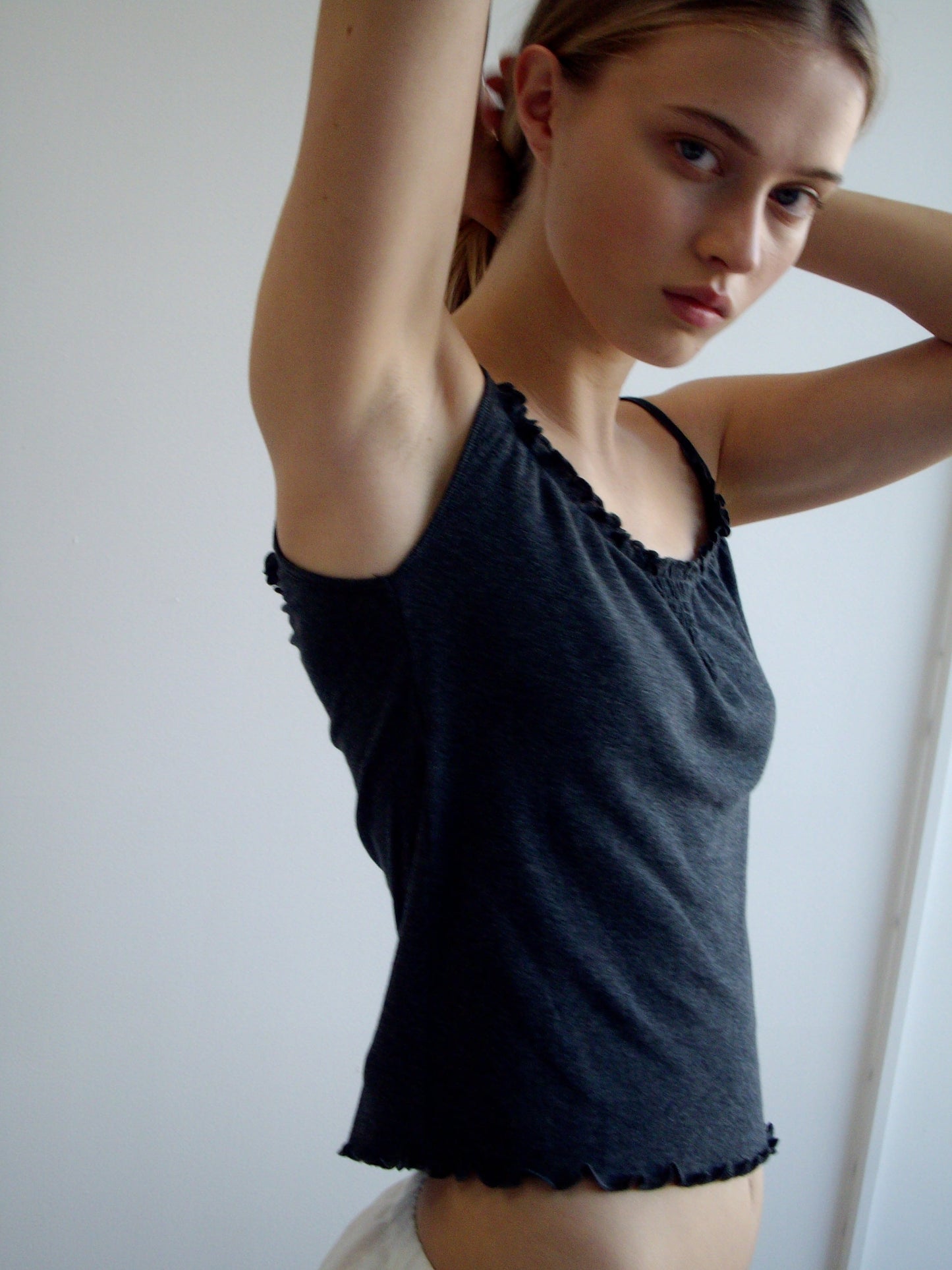 A young woman with light skin and straight light brown hair stands against a white background, wearing the Wool Blend Flounce Tank - Grey. She raises one arm to adjust her hair and gazes at the camera with a neutral expression.