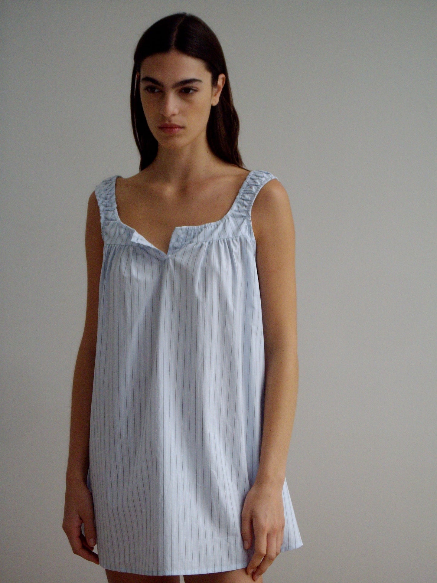 A woman with long dark hair stands against a plain background, wearing Deiji Studios’ Yoke Mini Dress in Mineral Stripe—an organic cotton mini dress featuring subtle vertical stripes and a square neckline. She has a neutral expression.