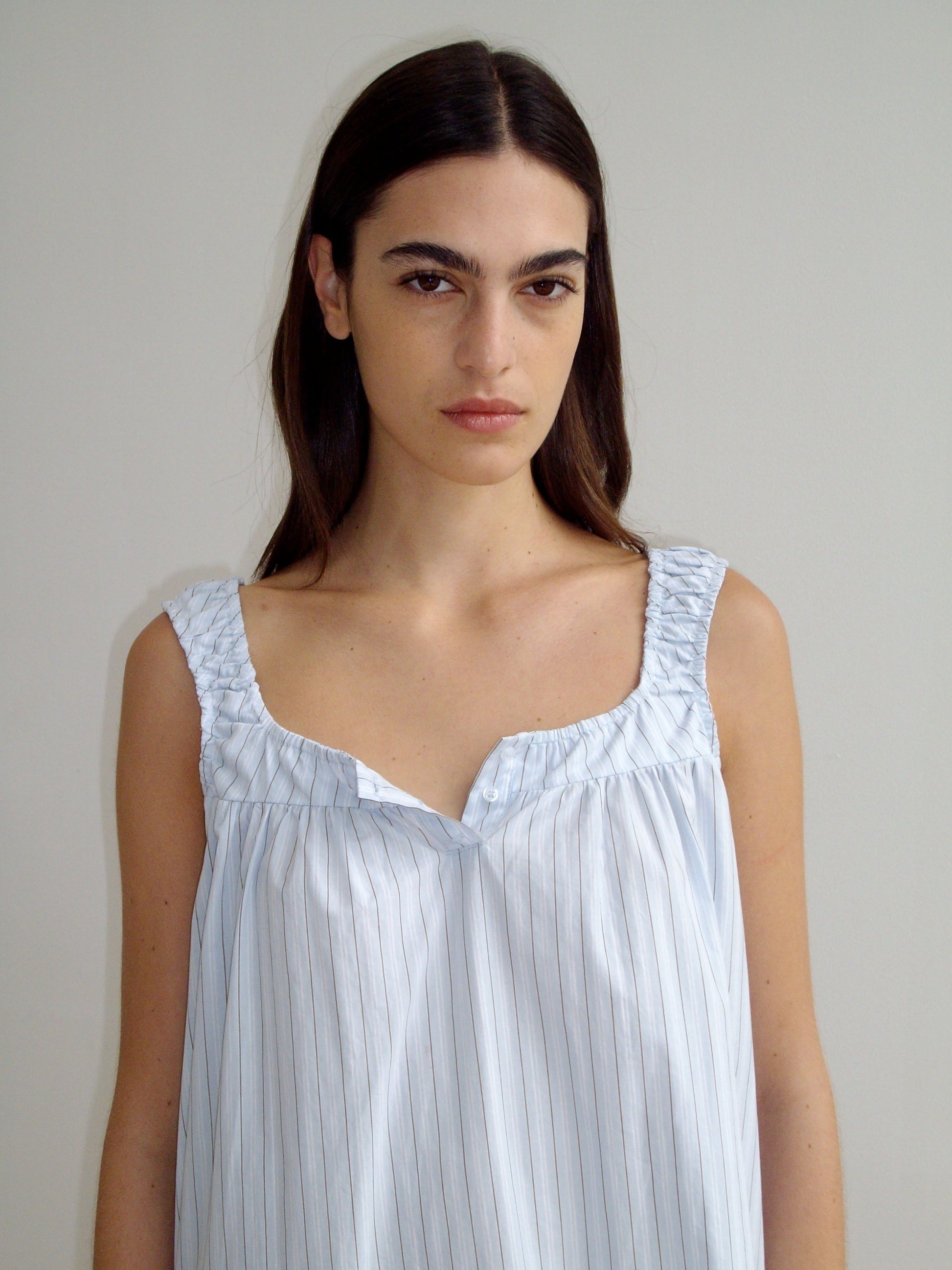 A woman with long brown hair stands against a plain light wall, wearing the Yoke Mini Dress in Mineral Stripe—a sleeveless, light blue and white striped organic cotton dress with ruched shoulder details. She looks directly at the camera with a neutral expression.