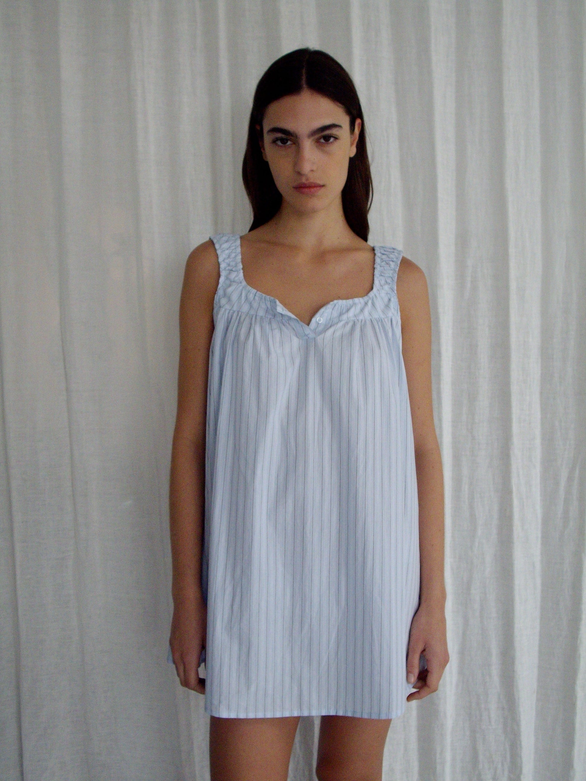 A woman with long dark hair stands before white curtains, wearing the Yoke Mini Dress - Mineral Stripe by Deiji Studios, a light blue, sleeveless, loose-fit mini dress in organic cotton with vertical stripes. She faces forward neutrally.