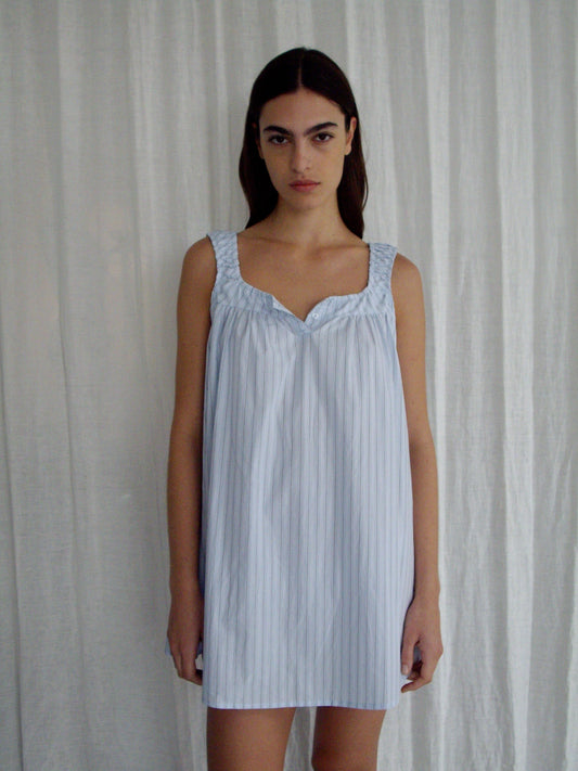 A woman with long dark hair stands before white curtains, wearing the Yoke Mini Dress - Mineral Stripe by Deiji Studios, a light blue, sleeveless, loose-fit mini dress in organic cotton with vertical stripes. She faces forward neutrally.