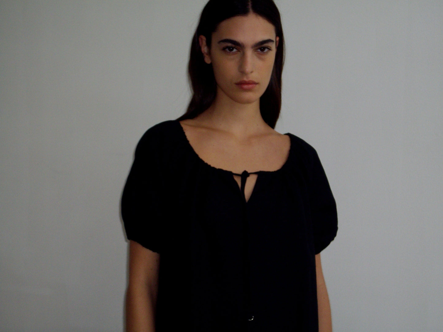 A person with long dark hair wears the Smock Dress - Black by Deiji Studios, featuring short sleeves and a front tie, standing against a plain light background and looking directly at the camera with a neutral expression.