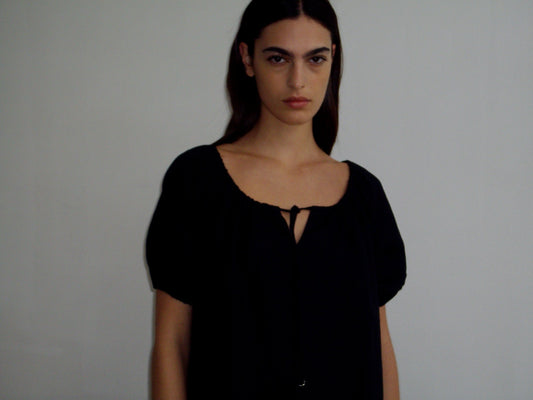 A person with long dark hair wears the Smock Dress - Black by Deiji Studios, featuring short sleeves and a front tie, standing against a plain light background and looking directly at the camera with a neutral expression.