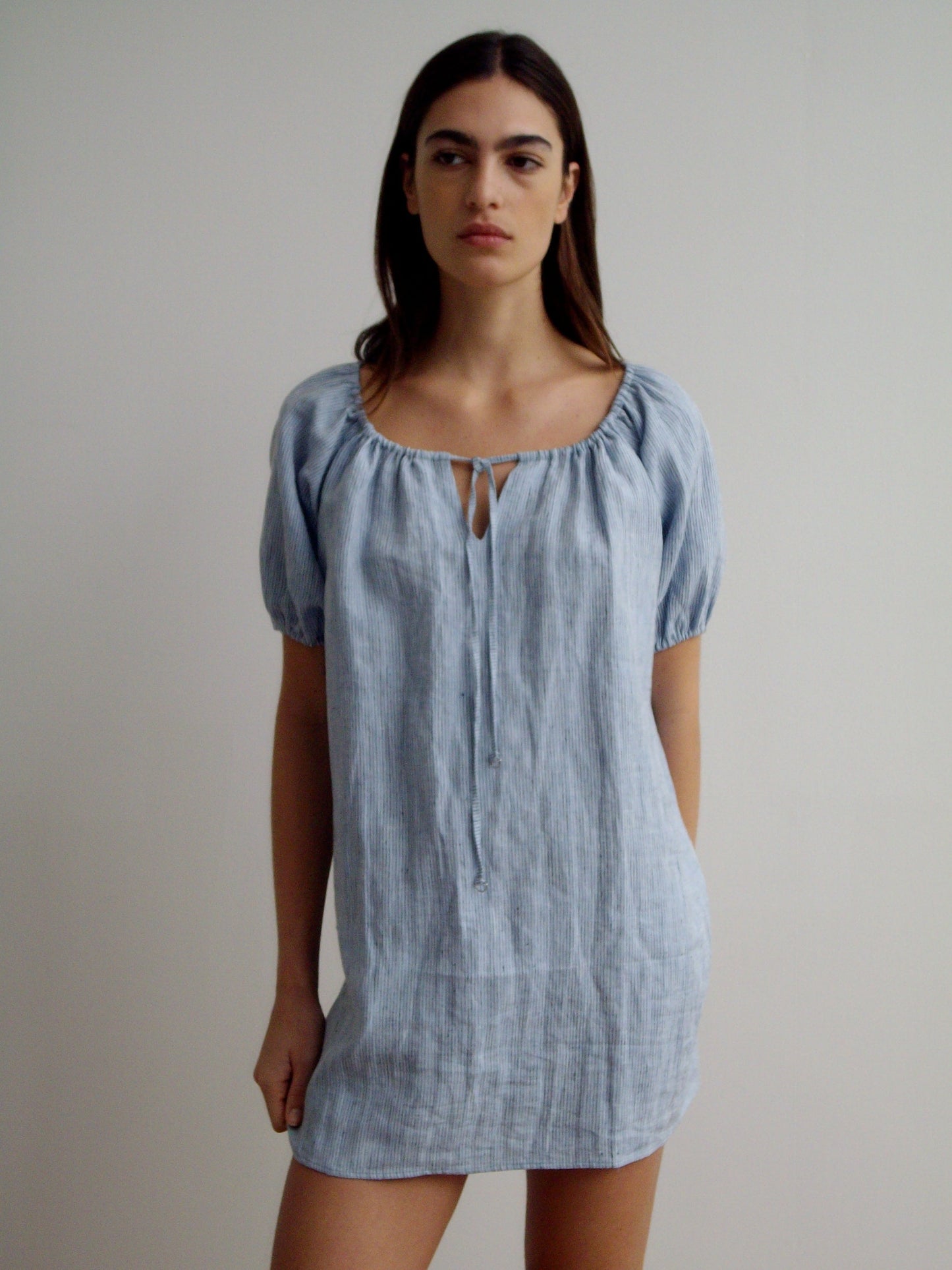 A woman with long dark hair and a neutral expression stands against a plain light background, wearing the Smock Dress - Blue Stripe with short puff sleeves, a round neckline, and a small keyhole detail.