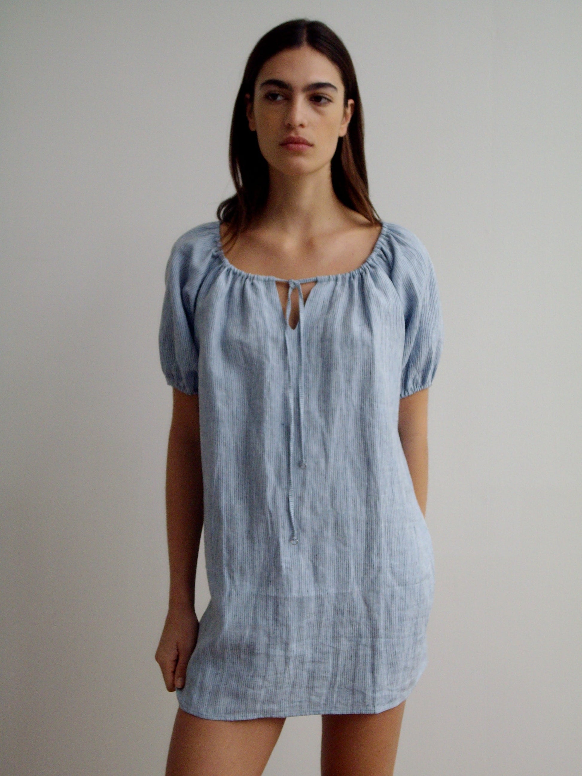 A woman with long dark hair and a neutral expression stands against a plain light background, wearing the Smock Dress - Blue Stripe with short puff sleeves, a round neckline, and a small keyhole detail.