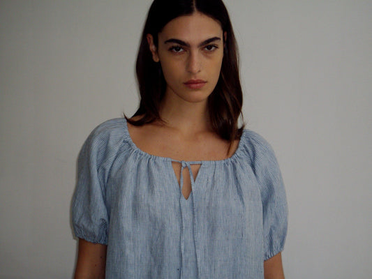 A person with long brown hair wears the Smock Dress - Blue Stripe and stands against a plain white background, looking directly at the camera with a neutral expression.