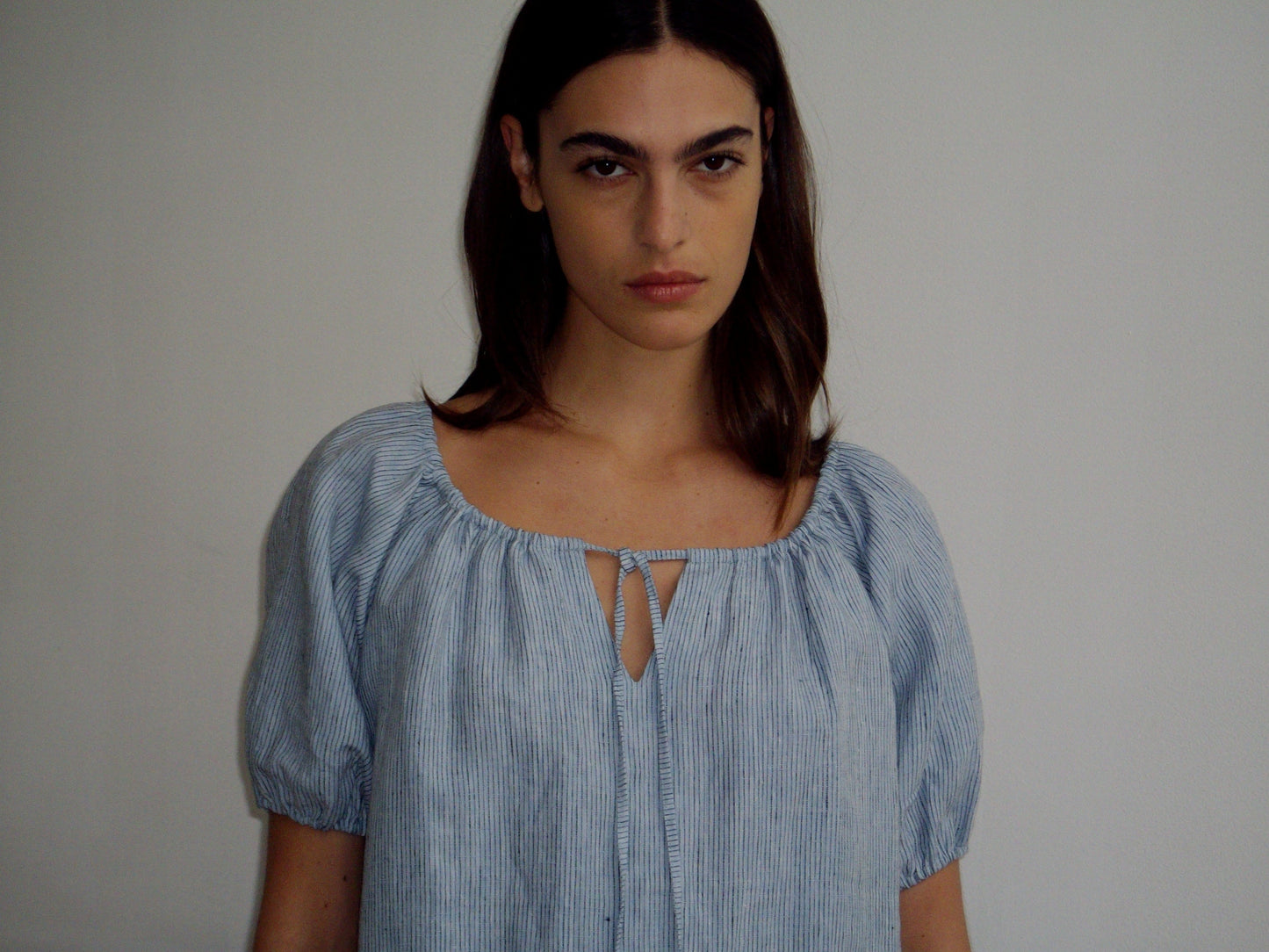 A woman with long brown hair stands against a plain light background, wearing a light blue, short-sleeved Smock Dress with a front tie, looking directly at the camera with a neutral expression.