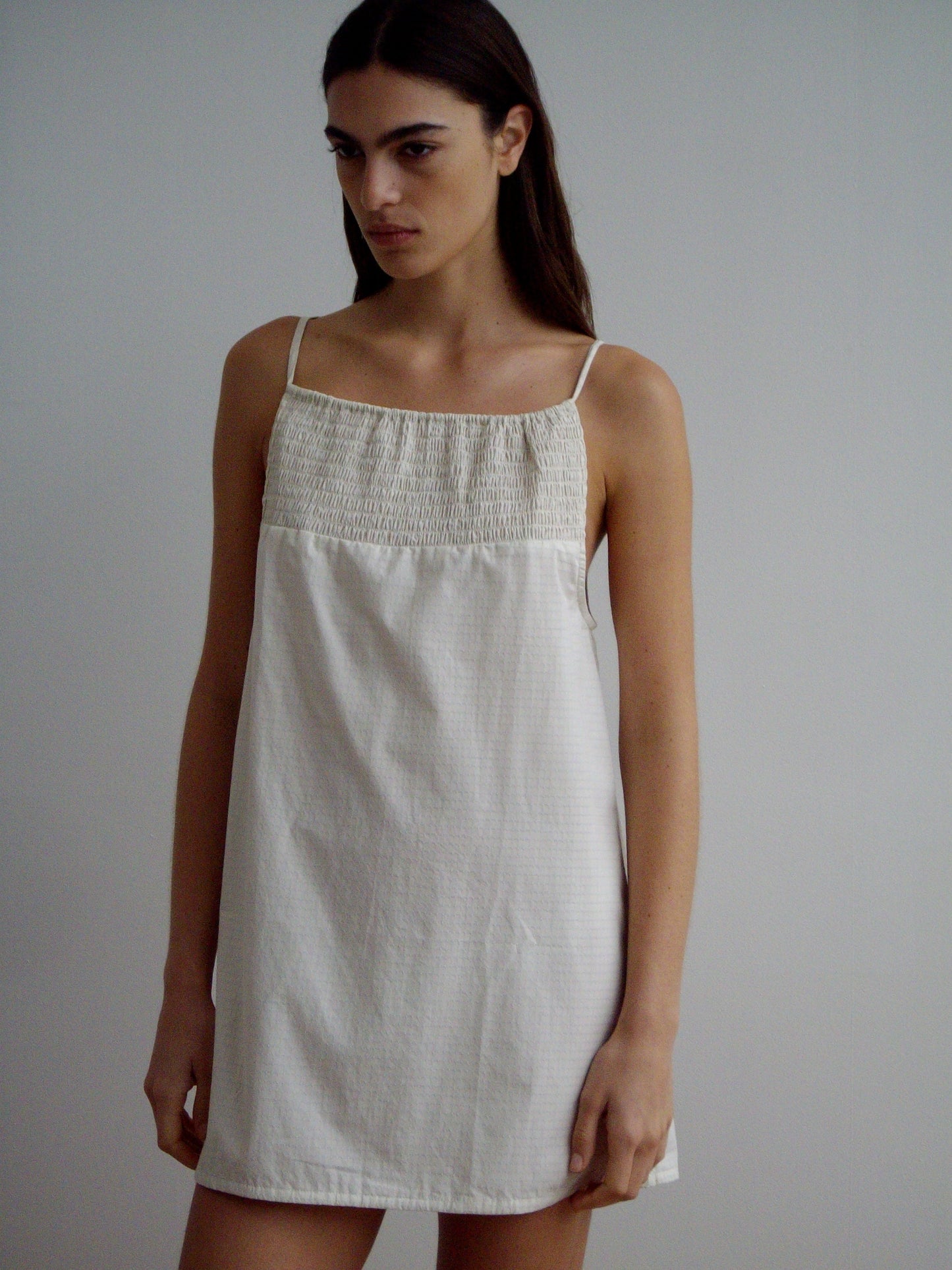 A woman with long brown hair wears the Shirring Mini Dress, a sleeveless, light-toned, loose-fitting style with thin straps. She stands against a plain light background, gazing slightly to the side with a neutral expression.