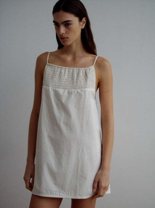 A woman with long dark hair models the Shirring Mini Dress, a sleeveless, light-toned dress featuring thin straps and a gathered top, set against a plain light background.