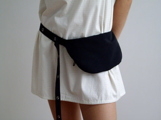 A person stands against a plain light background, wearing a white textured dress and the Snap Belt Bag - Black around their waist.