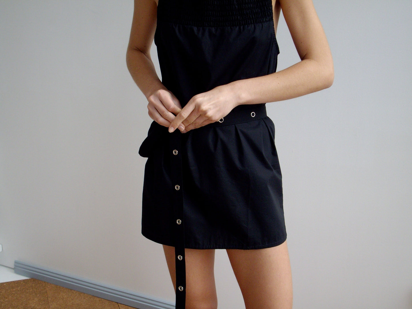 A person in a sleeveless black recycled polyester dress fastens a black belt with silver grommets at their waist and pairs it with the Snap Belt Bag - Black against a plain, light-colored background.