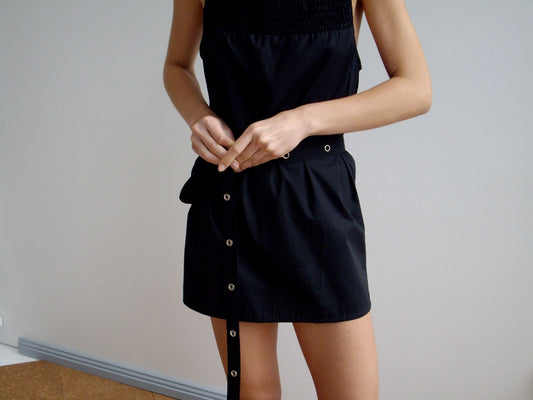 A person in a sleeveless black recycled polyester dress fastens a black belt with silver grommets at their waist and pairs it with the Snap Belt Bag - Black against a plain, light-colored background.
