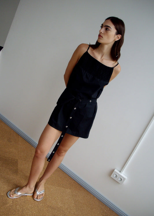 A woman wears the Deiji Studios Shirring Mini Dress in black with sandals, standing indoors on a brown floor against a plain white wall, showcasing the dress's minimal style.