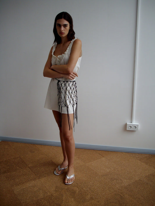 Indoors, a woman with crossed arms and a neutral expression wears a white sleeveless dress, silver sandals, and the Macrame Belt - Black around her waist. The plain background features a visible power outlet on the wall.