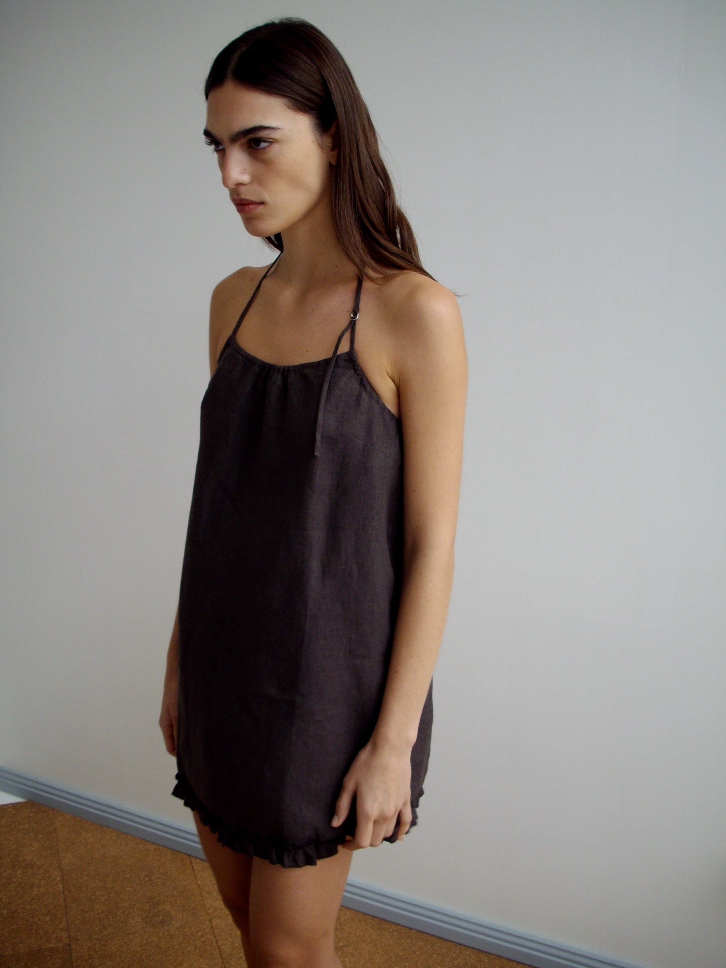 A woman with long brown hair stands indoors against a plain light wall, wearing the Flounce Halter Dress - Bark, a sleeveless, loose-fitting linen dress with thin straps and a ruffled hem, looking slightly to the side.