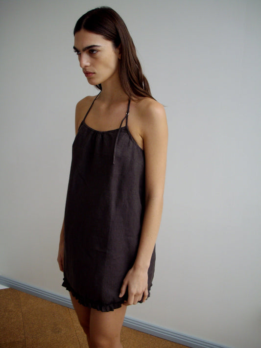 A woman with long brown hair stands indoors against a plain light wall, wearing the Flounce Halter Dress - Bark, a sleeveless, loose-fitting linen dress with thin straps and a ruffled hem, looking slightly to the side.
